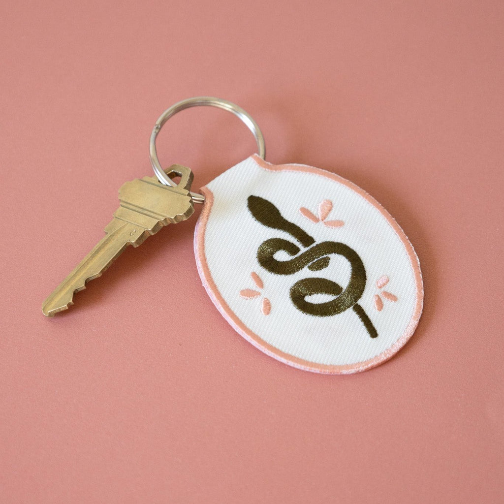 A white cotton twill keychain with an embroidered snake design, displayed on a pink background.