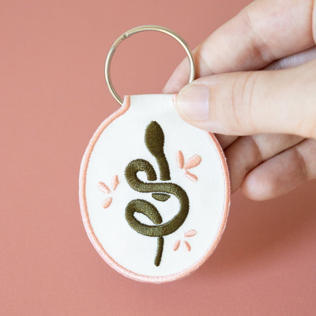 A hand holding a white cotton twill keychain with an embroidered snake design, displayed on a pink background.