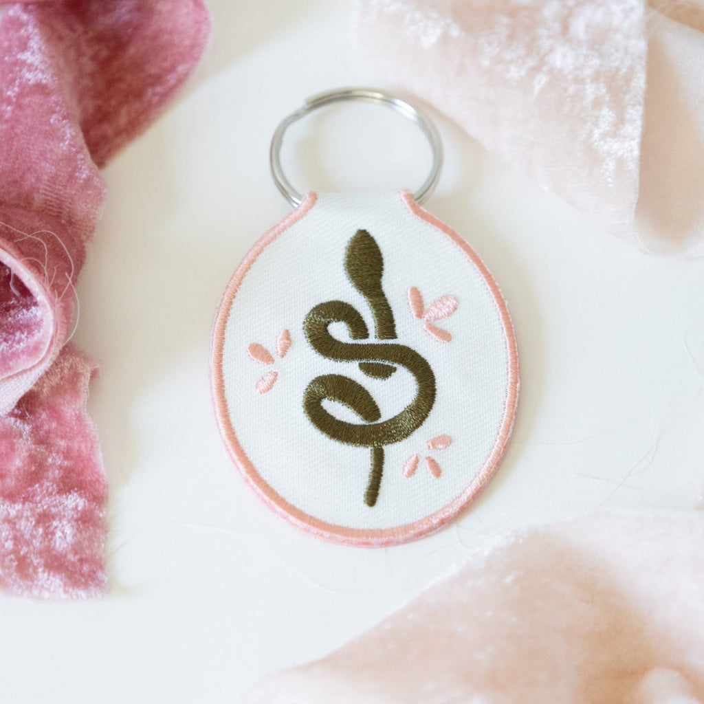 A white cotton twill keychain with an embroidered snake design, displayed on a white background with pink ribbons.