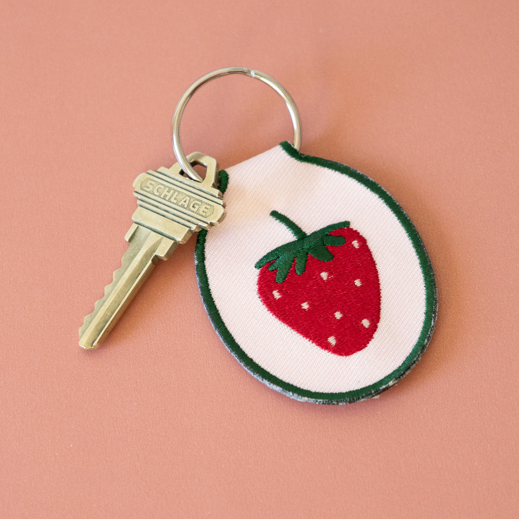 A light pink keychain with an embroidered strawberry design, displayed on a pink background.