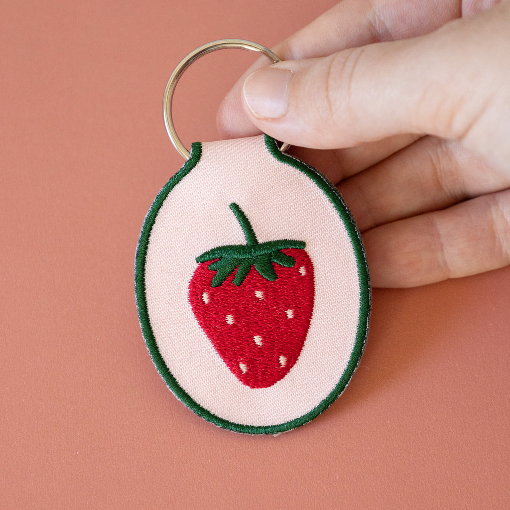 A hand holding a light pink keychain with an embroidered strawberry design, displayed on a pink background.