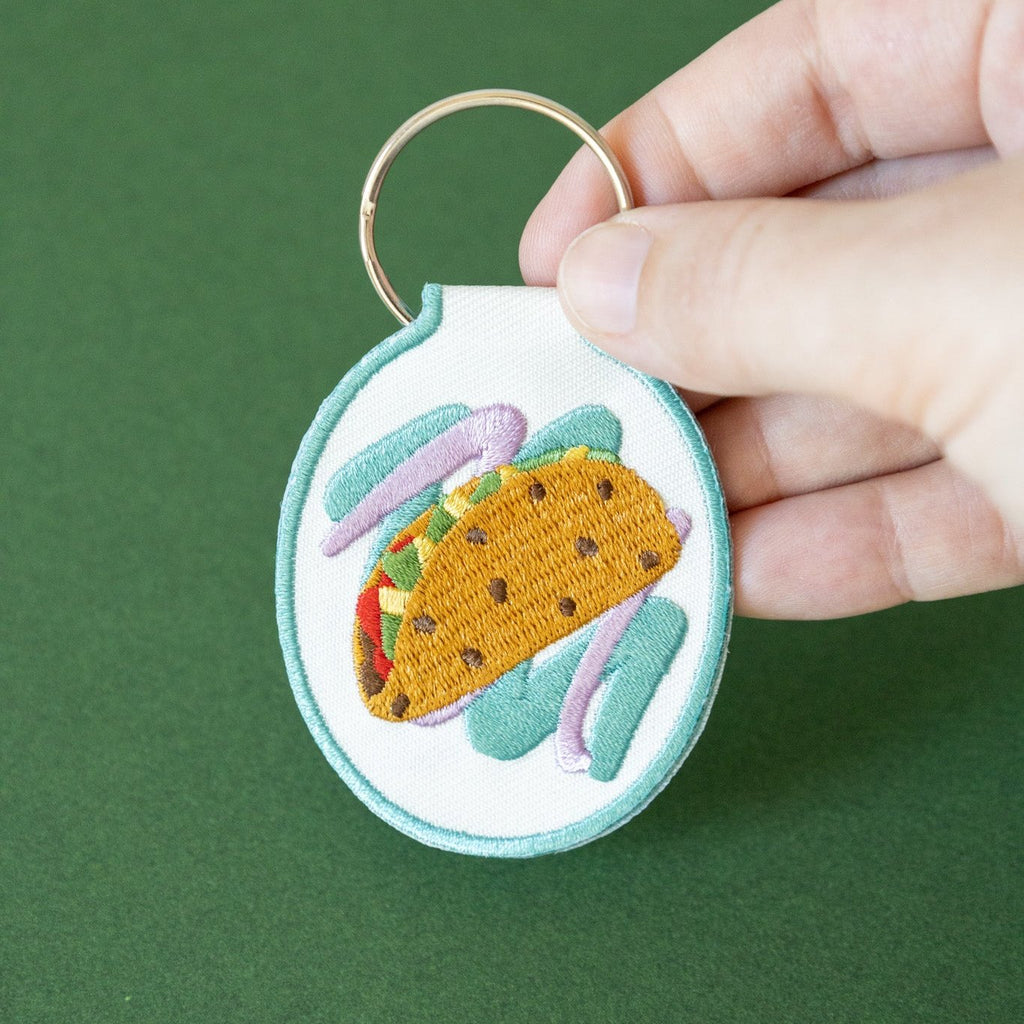 A hand holding an embroidered keychain with a taco design on white cotton twill, displayed against a green background.
