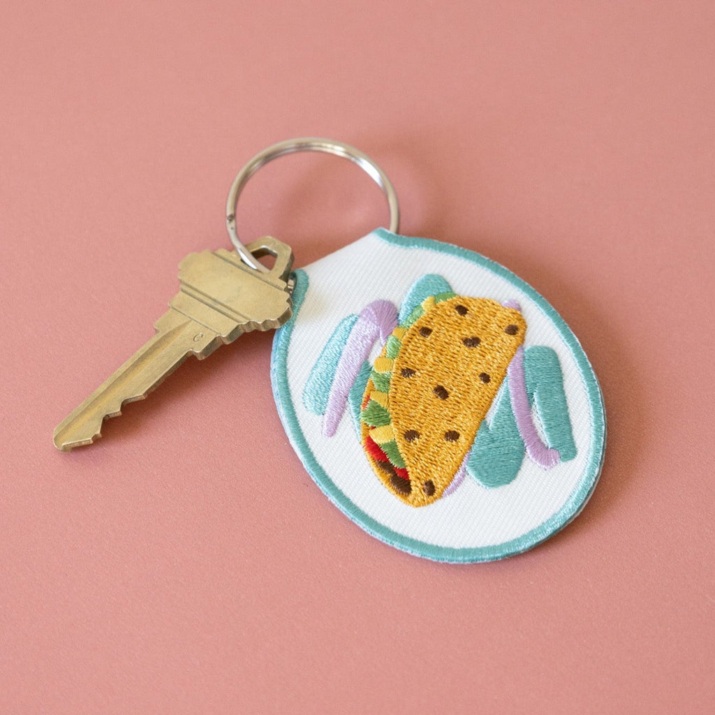 Embroidered keychain with a taco design on white cotton twill, displayed against a pink background.