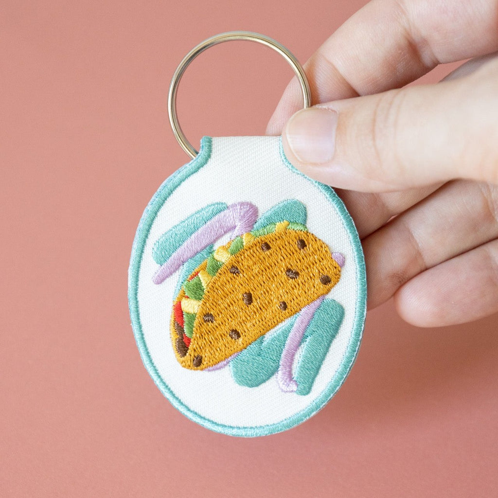 A hand holding an embroidered keychain with a taco design on white cotton twill, displayed against a pink background.