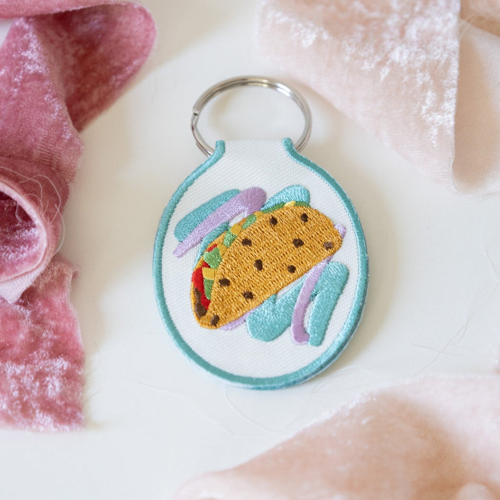 Embroidered keychain with a taco design on white cotton twill, displayed against a white background with pink ribbons.