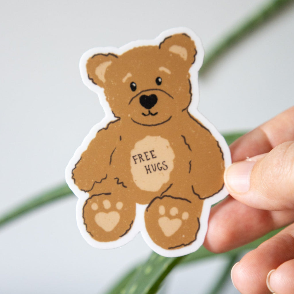 Sticker of a bear with 'Free Hugs' text held by a hand against a plant background