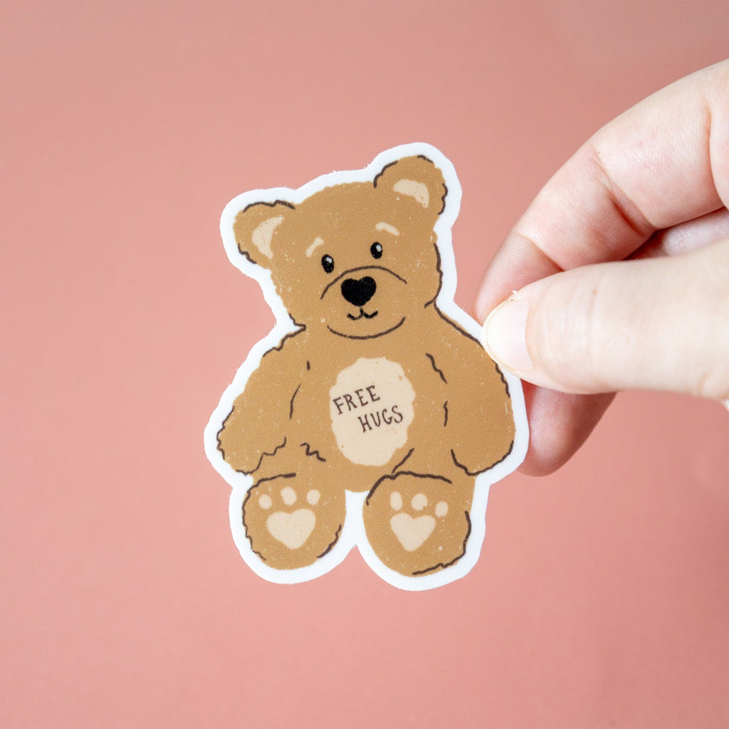 Sticker of a bear with 'Free Hugs' text held by a hand against a pink background