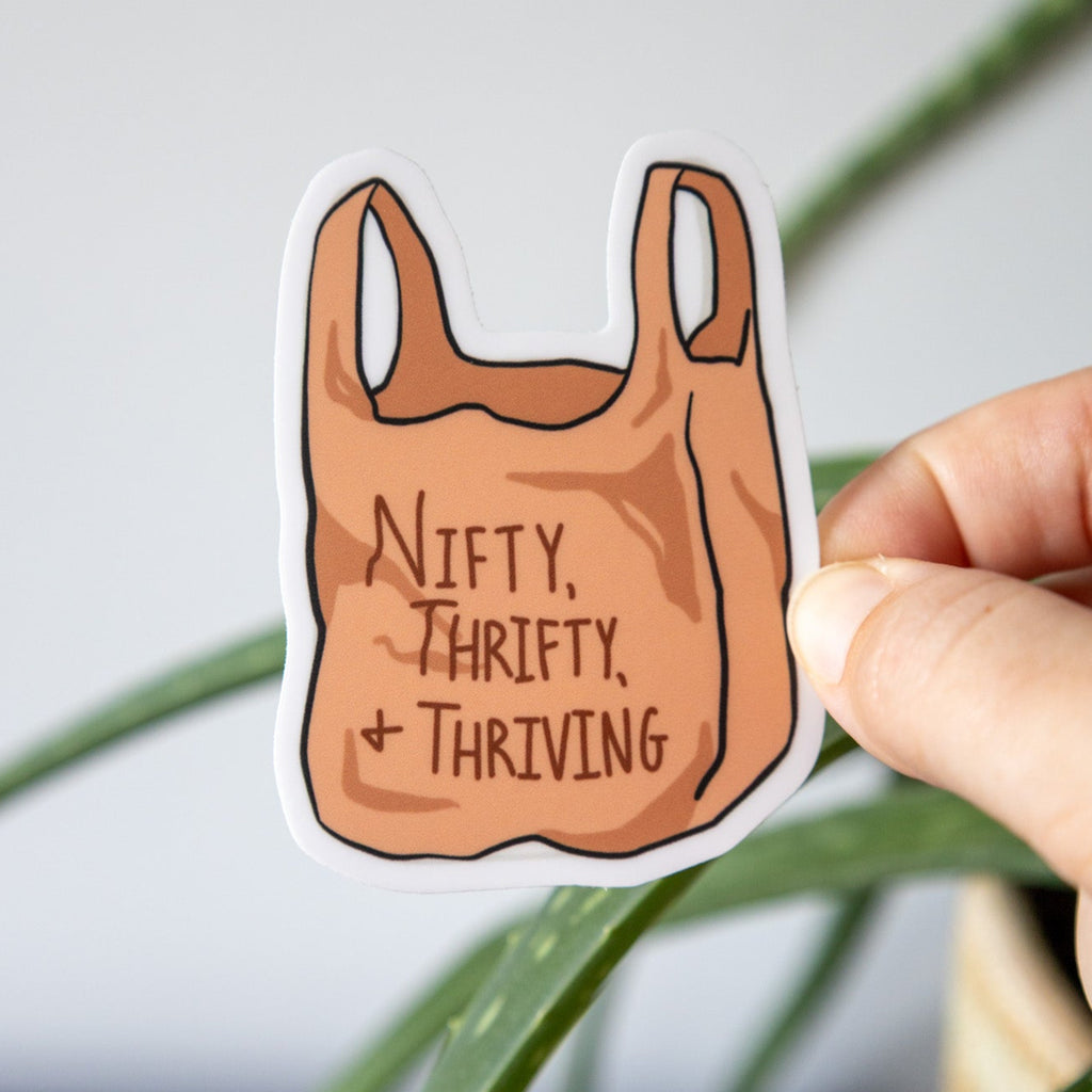 sticker of a shopping bag that says "nifty, thrifty, and thriving" held by a hand with a plant in the background