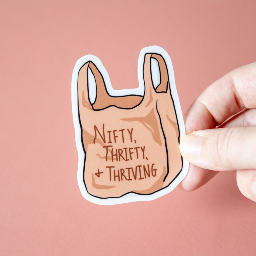 Sticker of a shopping bag with text 'Nifty, Thrifty, & Thriving' held by a hand against a pink background