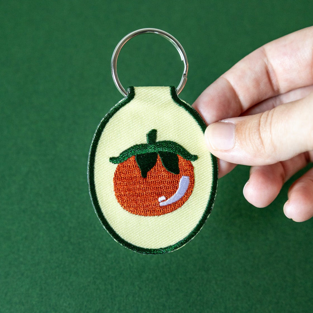 Hand holding a keychain with an embroidered tomato design on butter yellow fabric against a green background.