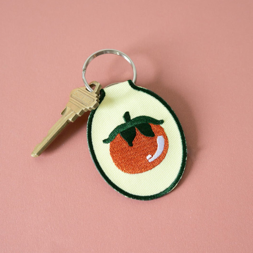 Embroidered keychain with tomato design on butter yellow fabric against a pink background.
