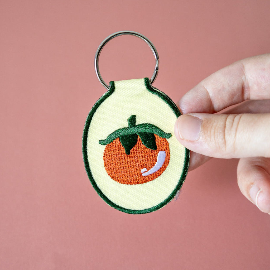 Hand holding a keychain with an embroidered tomato design on butter yellow fabric against a pink background.