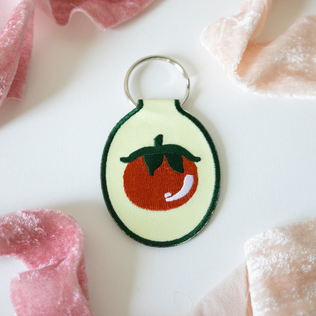 Embroidered keychain with a tomato design on butter yellow fabric sitting on a white background with pink ribbons.