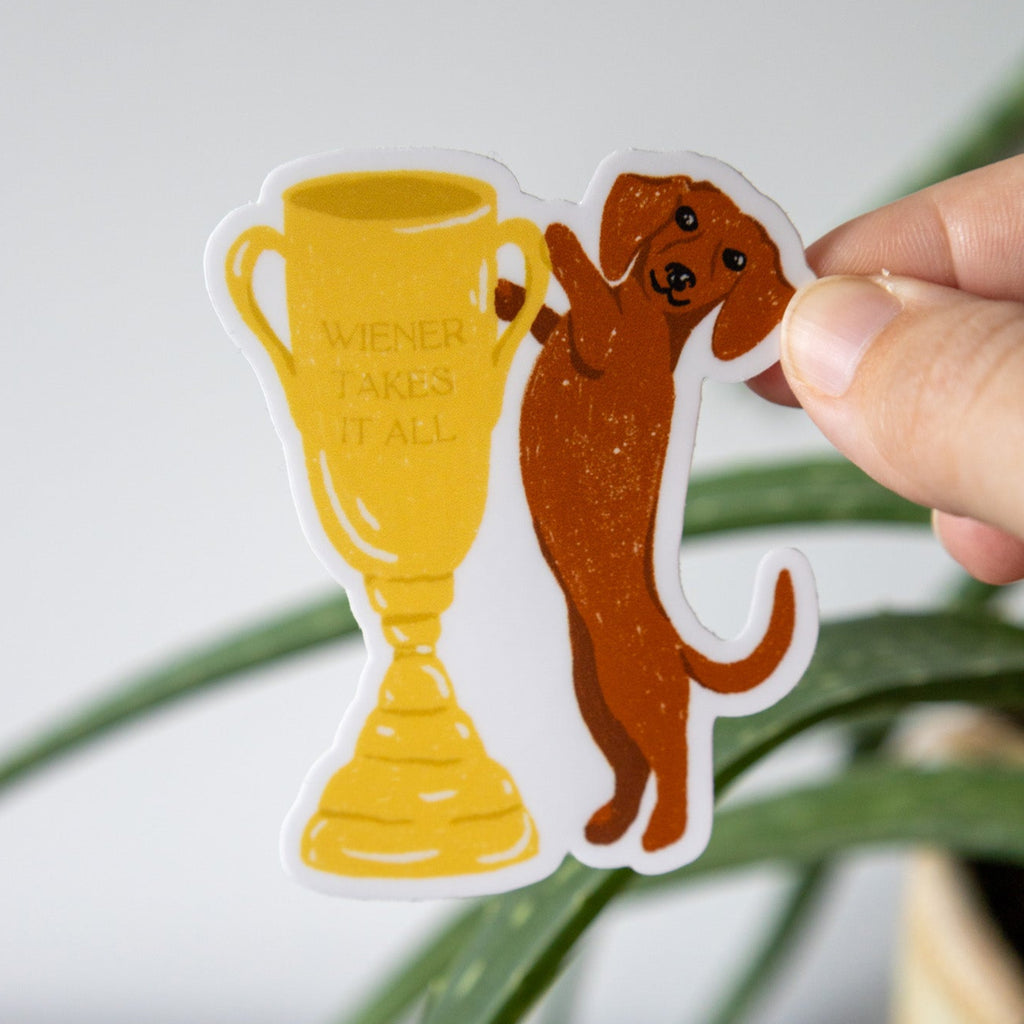 Hand holding a sticker of a dog next to a trophy that reads "wiener takes it all" against a wall with a plant in the background