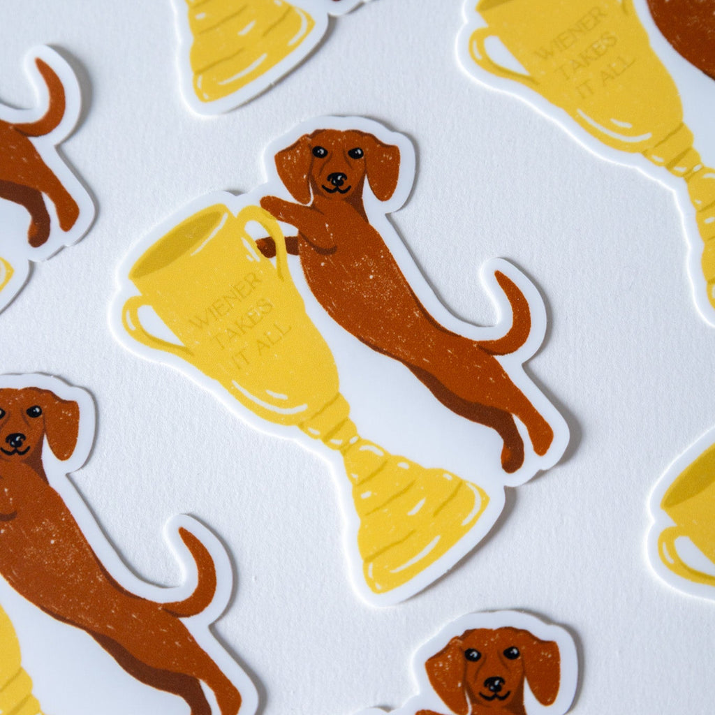 Stickers of a dog next to a trophy that reads "wiener takes it all" on a white background