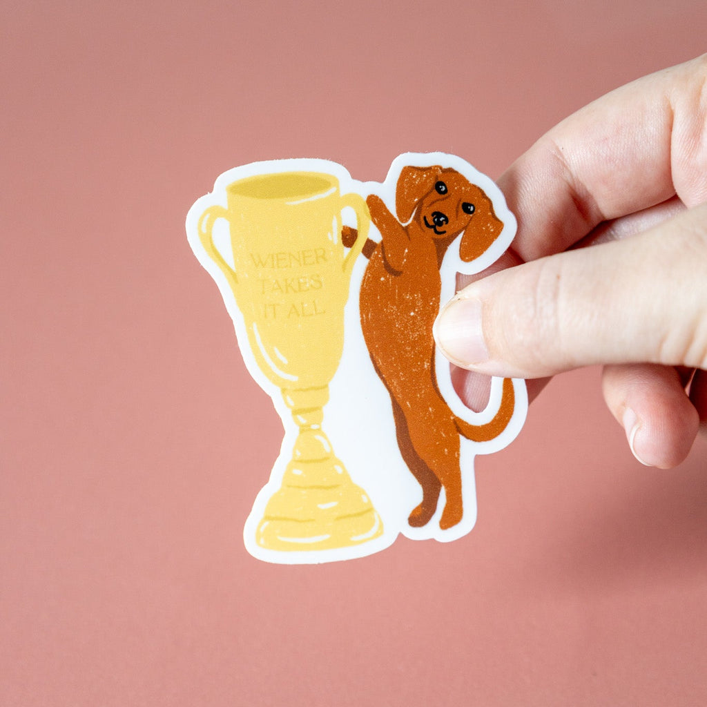 Hand holding a sticker of a dog next to a trophy that reads "wiener takes it all" on a pink background