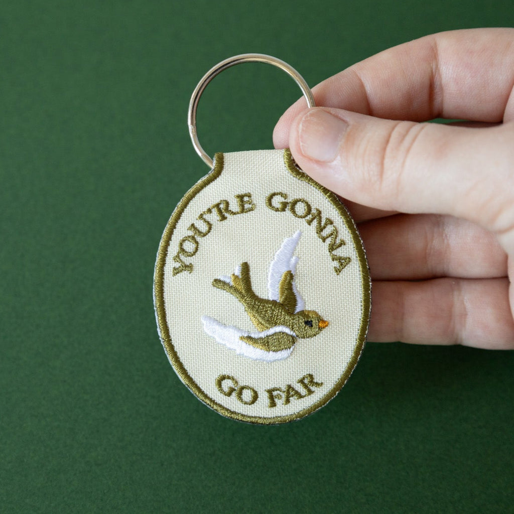 A hand holding a beige keychain with the embroidered text 'YOU'RE GONNA GO FAR' and an embroidered bird illustration on it, displayed against a green background.