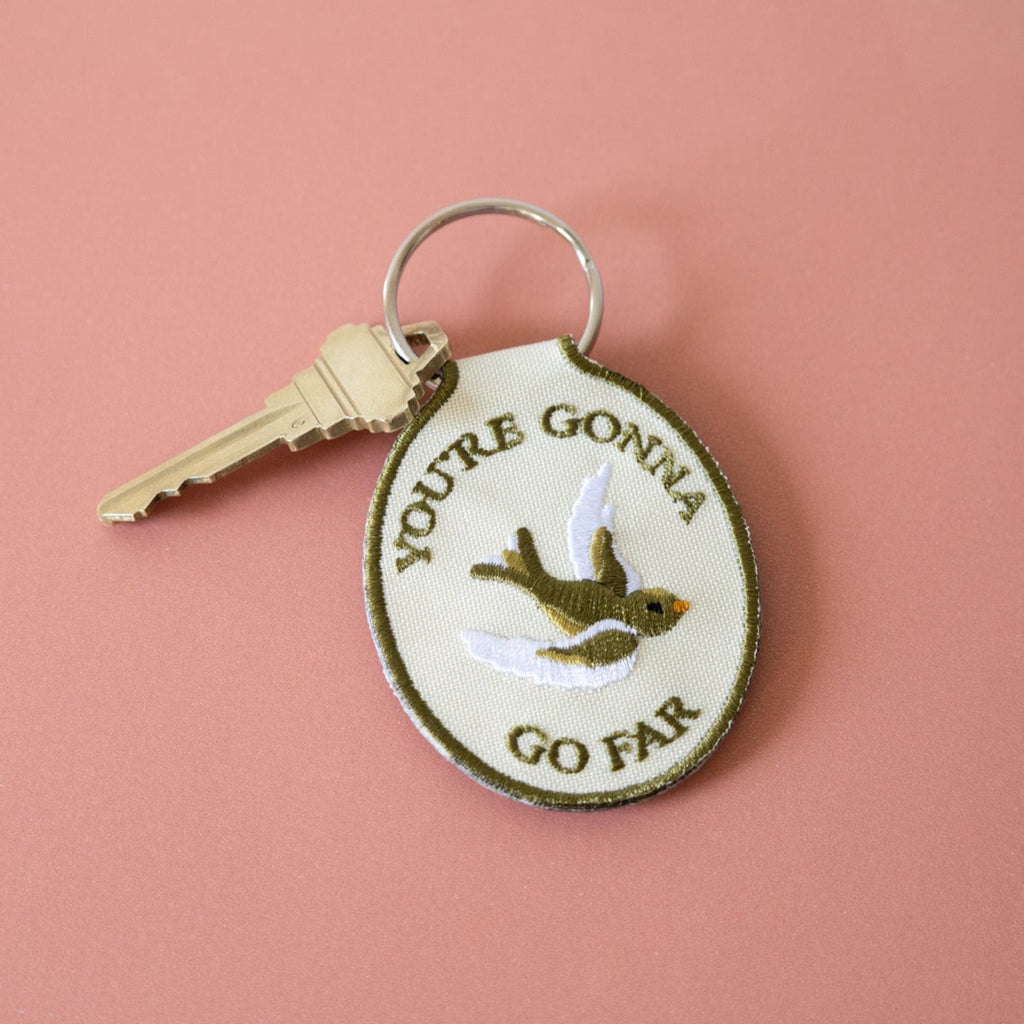 A beige keychain with the embroidered text 'YOU'RE GONNA GO FAR' and an embroidered bird illustration on it, displayed against a pink background.