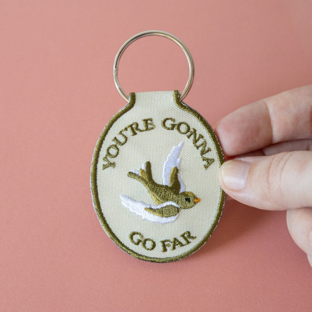 A hand holding a beige keychain with the embroidered text 'YOU'RE GONNA GO FAR' and an embroidered bird illustration on it, displayed against a pink background.
