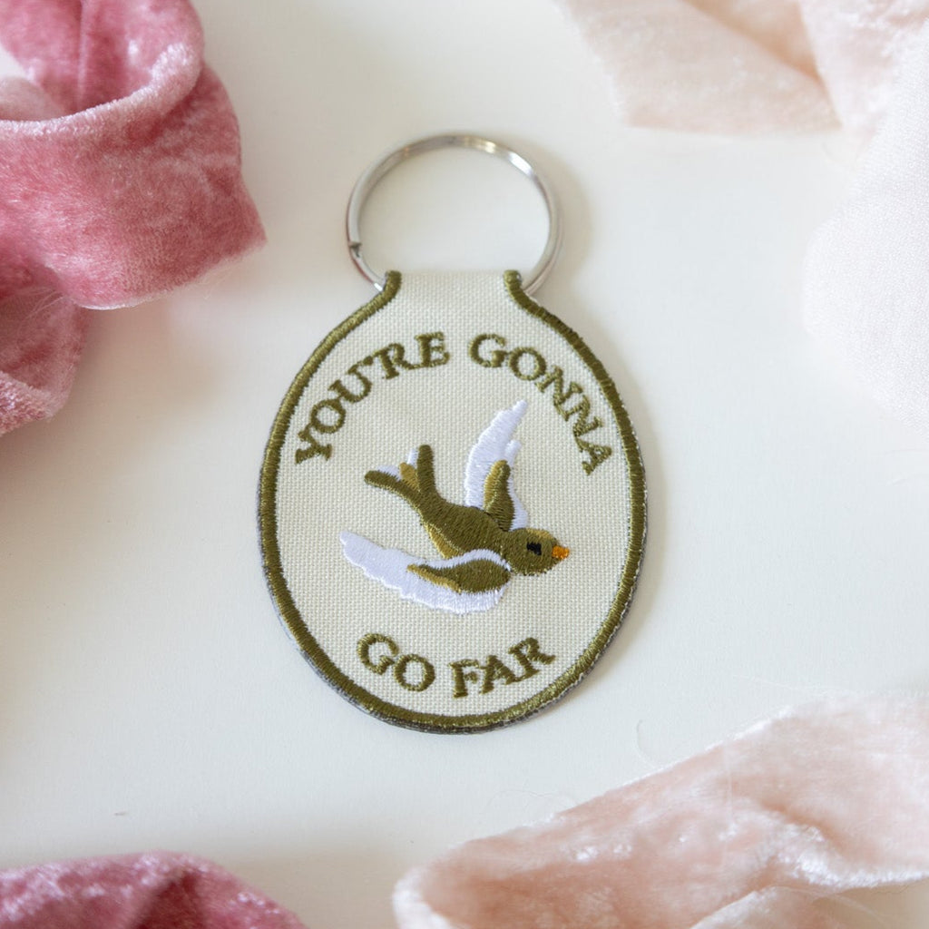 A beige keychain with the embroidered text 'YOU'RE GONNA GO FAR' and an embroidered bird illustration on it, displayed against a white background with pink ribbons.